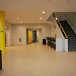 Entryway to the indoor storage units with a carpeted staircase to the right and elevators and a yellow pillar to the left. At the back of the entryway is a doorway leading to indoor storage units